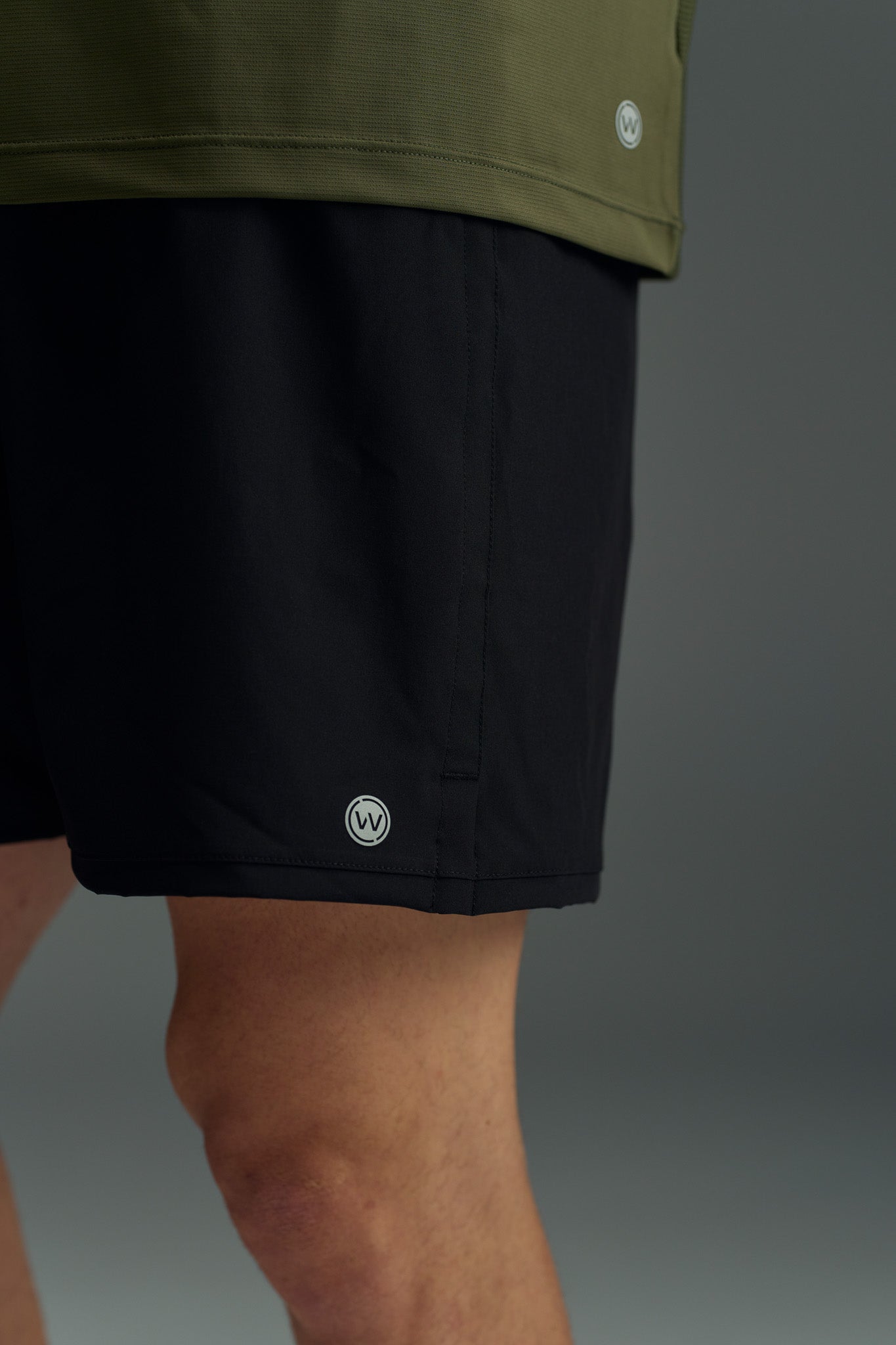 A close-up shows a person wearing the WearEver Cadence Short in black (5-inch inseam) and an olive green top, both by WearEver with small circular logos near the hem. The background is plain and neutral.