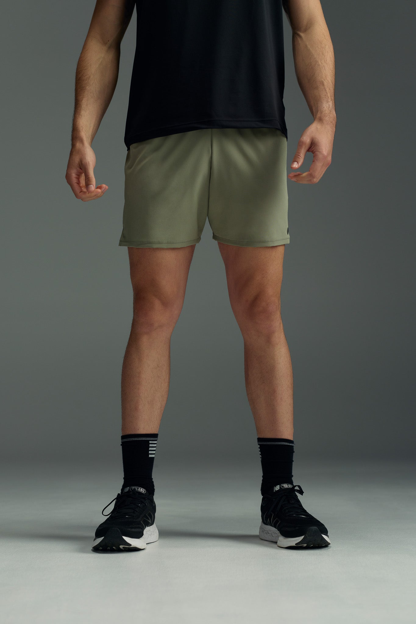 A person stands with arms relaxed at their sides, wearing a black T-shirt and WearEver Cadence Short lightweight running shorts with antibacterial treatment, paired with black socks and athletic shoes, against a plain gray background. Face not visible.