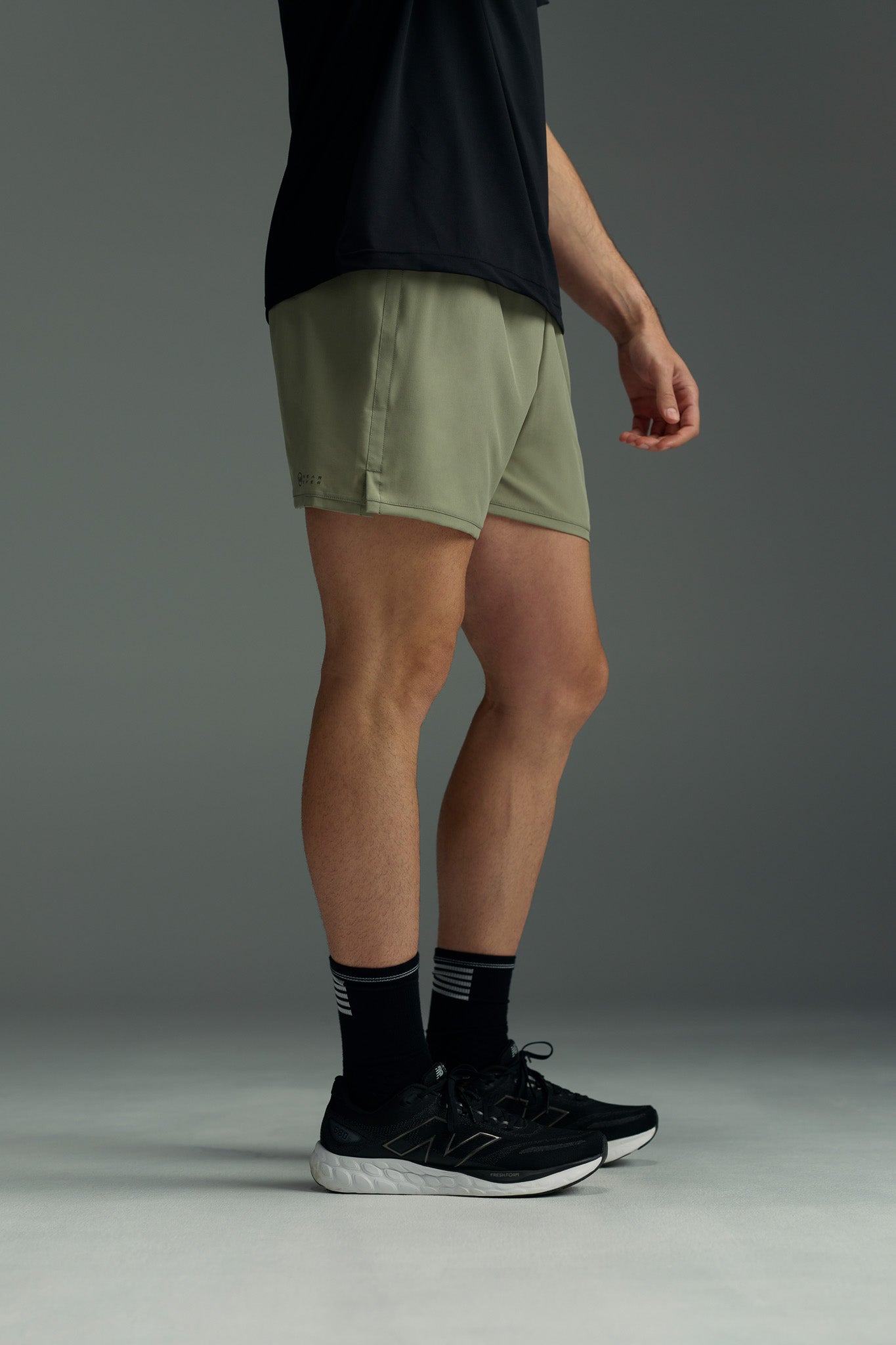 A person stands sideways in a black shirt, WearEver Cadence Short (5" inseam, olive green), black socks with white stripes, and black running shoes against a plain gray background.