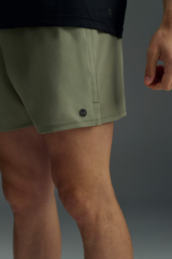 A close-up shows a person in olive green WearEver Cadence Shorts (5" inseam) and a black shirt, right arm relaxed against a plain grey backdrop. The lightweight, breathable WearEver fabric delivers comfort all day.