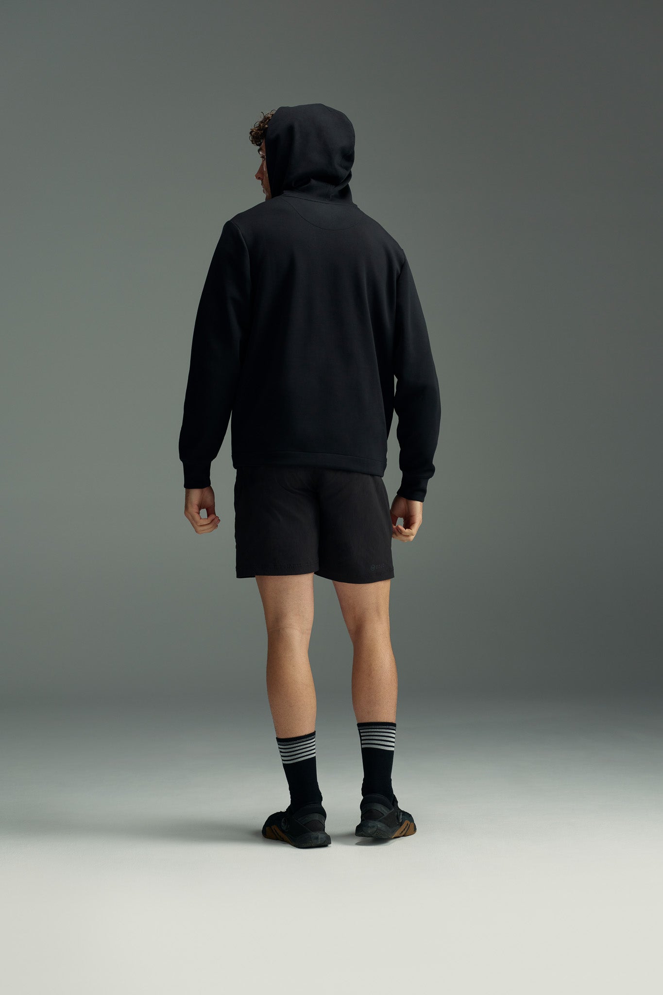 A person in a black hoodie, black socks with white stripes, black shoes, and WearEver Agile Short by WearEver—7-inch inseam lightweight stretch shorts with an elastic waistband—stands facing away against a plain gray background.