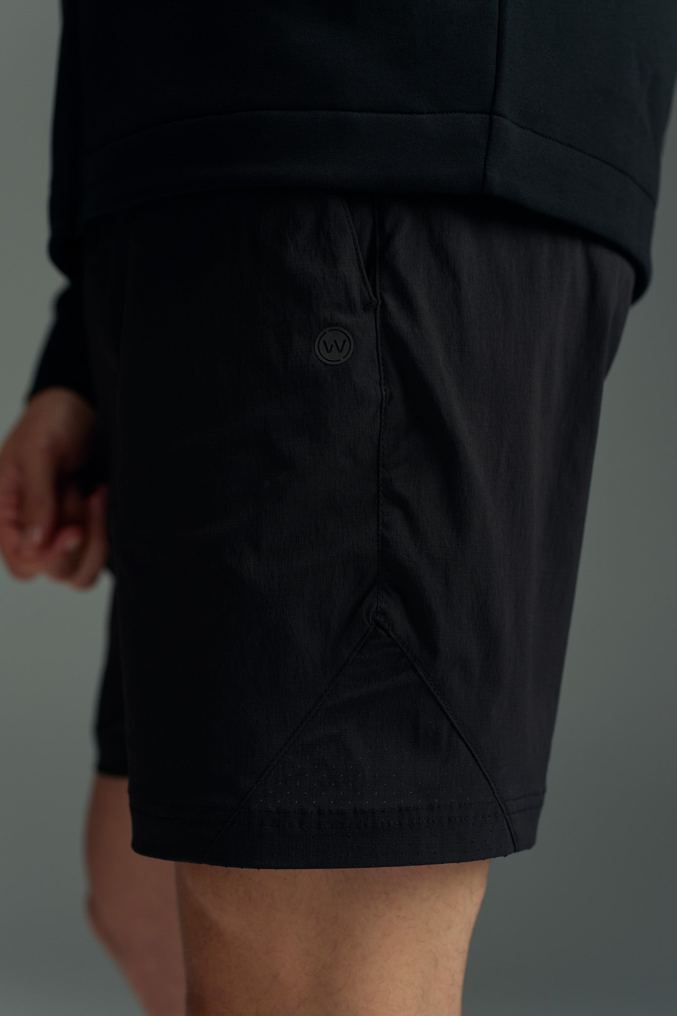 A person wears WearEver Agile Shorts and a black top, standing with one arm relaxed. The focus is on the lightweight, stretchy WearEver shorts, featuring a 7-inch inseam, textured fabric, elastic waistband, and small round logo.