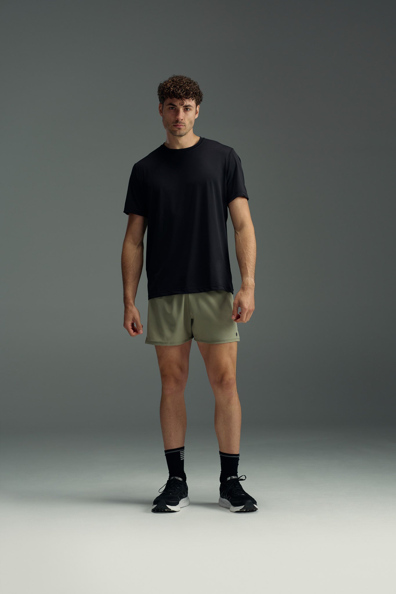 A man stands against a gray background, facing forward in a black t-shirt, black socks and sneakers, and WearEver Cadence Short—olive green, lightweight, breathable athletic shorts with a 5-inch inseam.