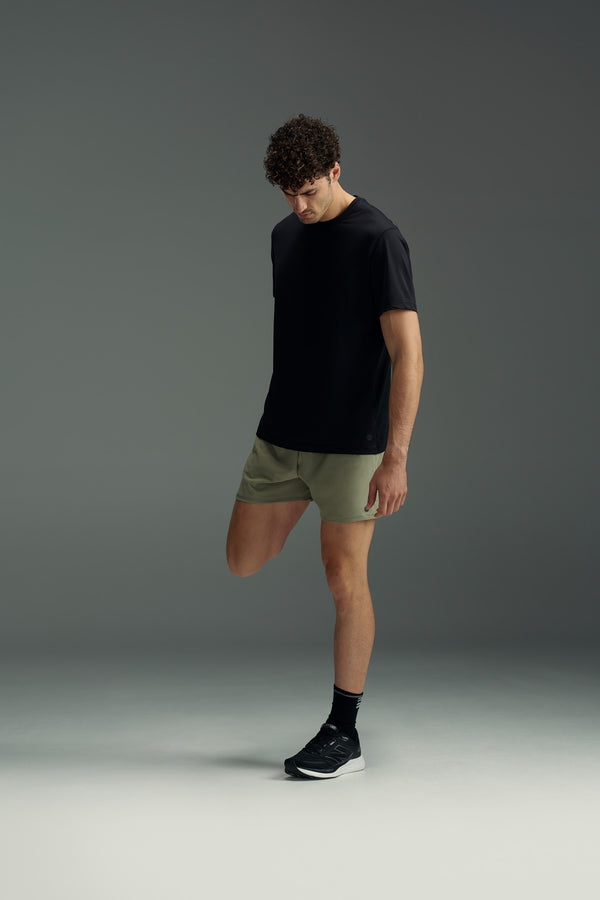 A man in a black T-shirt, WearEver Cadence Short with a 5-inch inseam, black socks, and sneakers stands on one leg holding his ankle, head down, against a plain gray background.