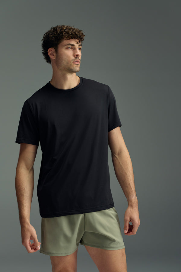A man with curly hair stands against a gray background, wearing the WearEver Pulse Tee in black and light olive green shorts from WearEver. He looks to his left with a neutral expression.