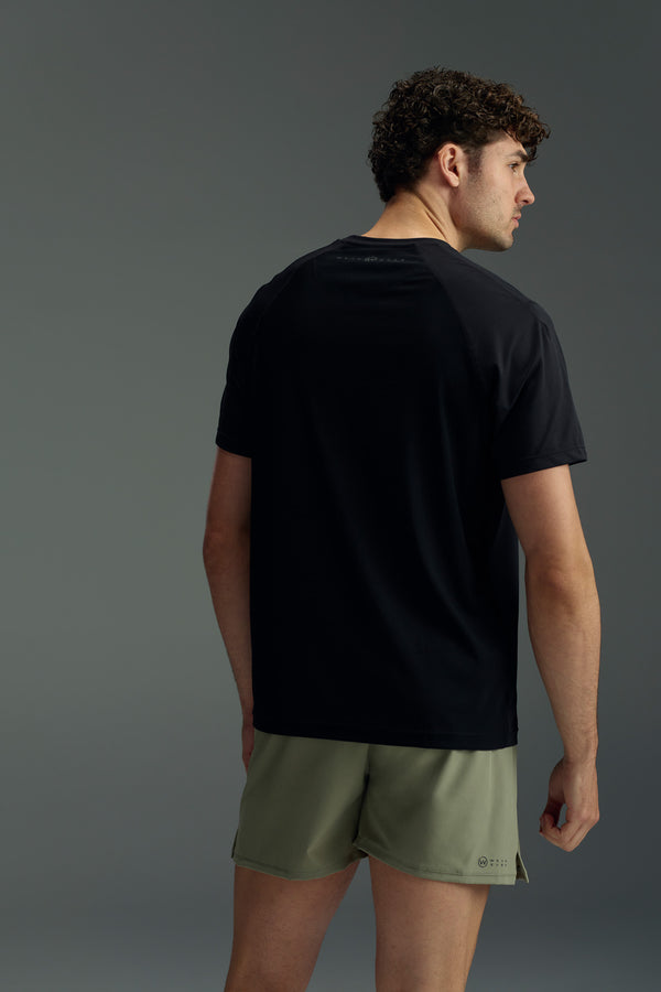 A man with short curly hair faces away, wearing the WearEver Pulse Tee—a black quick-dry performance shirt by WearEver—and olive green shorts against a plain gray background.