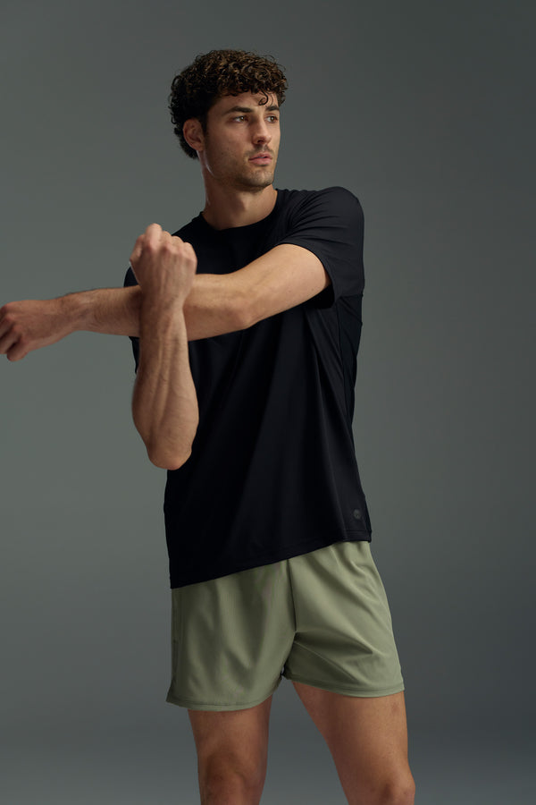 A man wearing the WearEver Pulse Tee in black stretches his arm across his chest while sporting olive green athletic shorts, posed against a plain gray background.