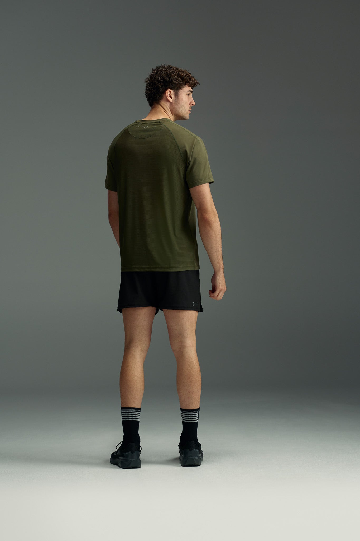 A man faces away, wearing the olive green WearEver Pulse Tee by WearEver, paired with black shorts, black socks with white stripes, and black shoes against a plain gray background.