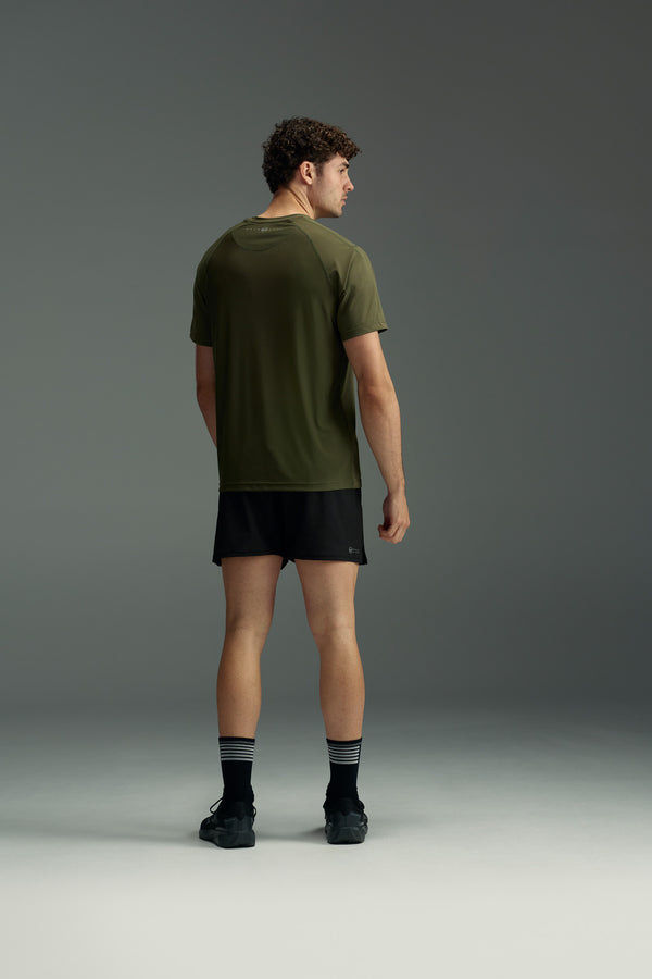 A man faces away, wearing the olive green WearEver Pulse Tee by WearEver, paired with black shorts, black socks with white stripes, and black shoes against a plain gray background.
