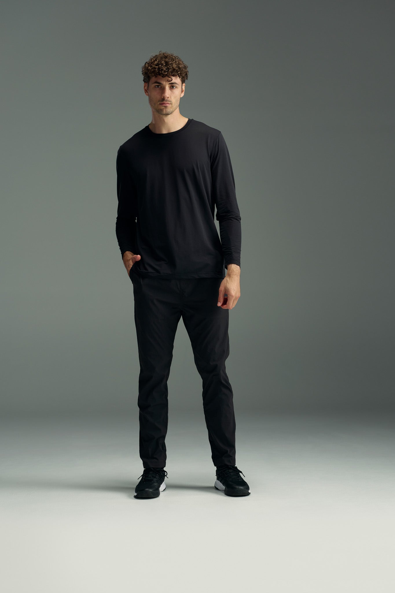 A man with curly hair stands against a gray background, wearing the WearEver Pulse Long Sleeve Tee by WearEver, paired with black pants and black sneakers, with one hand in his pocket.