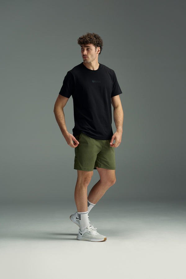 A man stands in a studio wearing the WearEver Outbound T-Shirt by WearEver, olive green shorts, white crew socks, and white sneakers. He looks to the side with one hand at his side and the other near his shorts pocket.