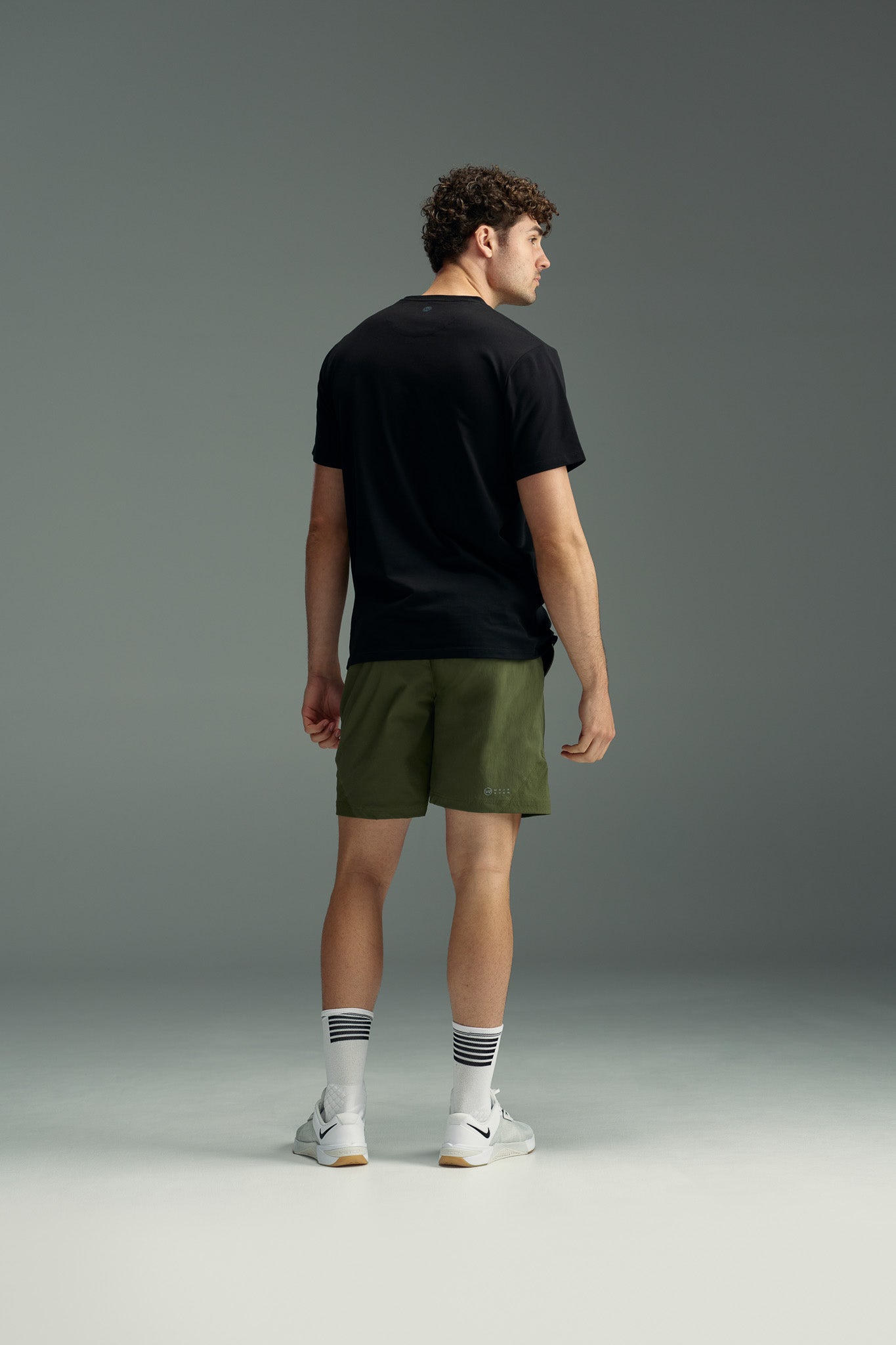 A man stands facing away, wearing a black T-shirt and olive green WearEver Agile Shorts with a 7-inch inseam, white socks featuring black stripes, and white sneakers, set against a plain gray background.