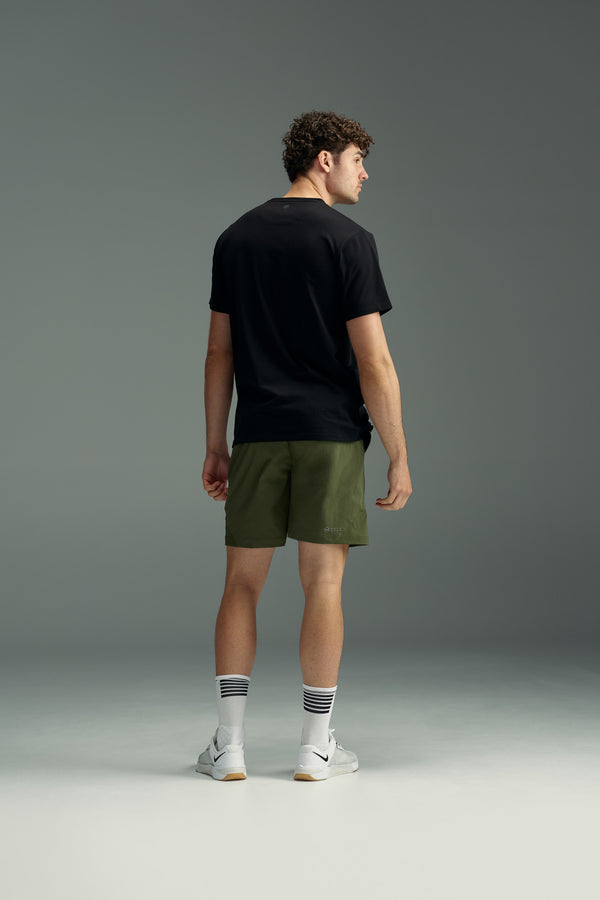 A man stands facing away, wearing a black T-shirt and olive green WearEver Agile Shorts with a 7-inch inseam, white socks featuring black stripes, and white sneakers, set against a plain gray background.