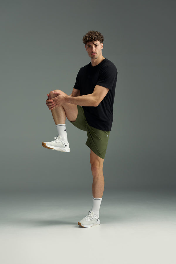A young man in athletic wear stretches his right leg, wearing a black t-shirt, WearEver Agile Short by WearEver with a 7-inch inseam, white socks, and white sneakers against a plain background.