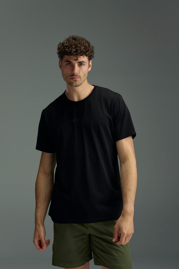 A man with curly hair wears the black WearEver Outbound T-Shirt and olive green shorts, standing against a plain gray background and looking directly at the camera with a neutral expression.