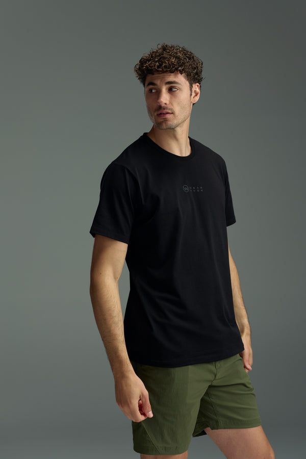 A man with curly hair wears a black WearEver Outbound T-Shirt and olive green shorts, standing against a gray background and looking to his left.