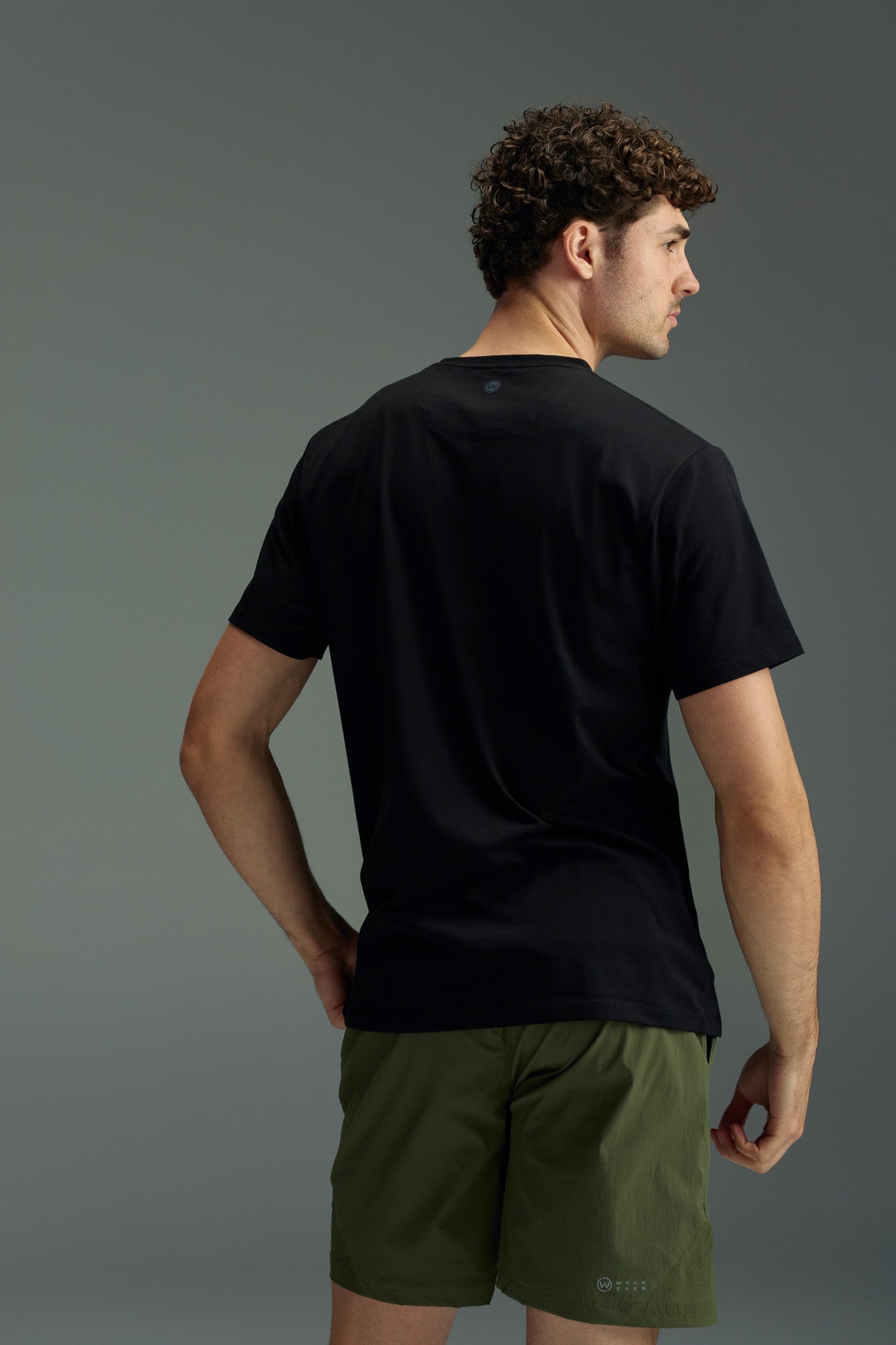 A man with curly hair faces away, wearing the WearEver Outbound T-Shirt by WearEver and olive green shorts against a plain gray background. His left hand is on his hip.