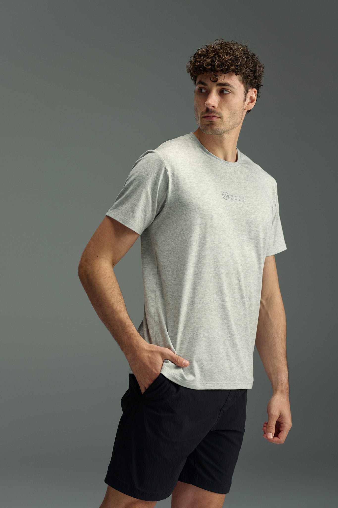 A man with curly hair wears the WearEver Outbound T-Shirt in light gray and black shorts, standing with one hand in his pocket and looking to the side against a plain gray background.