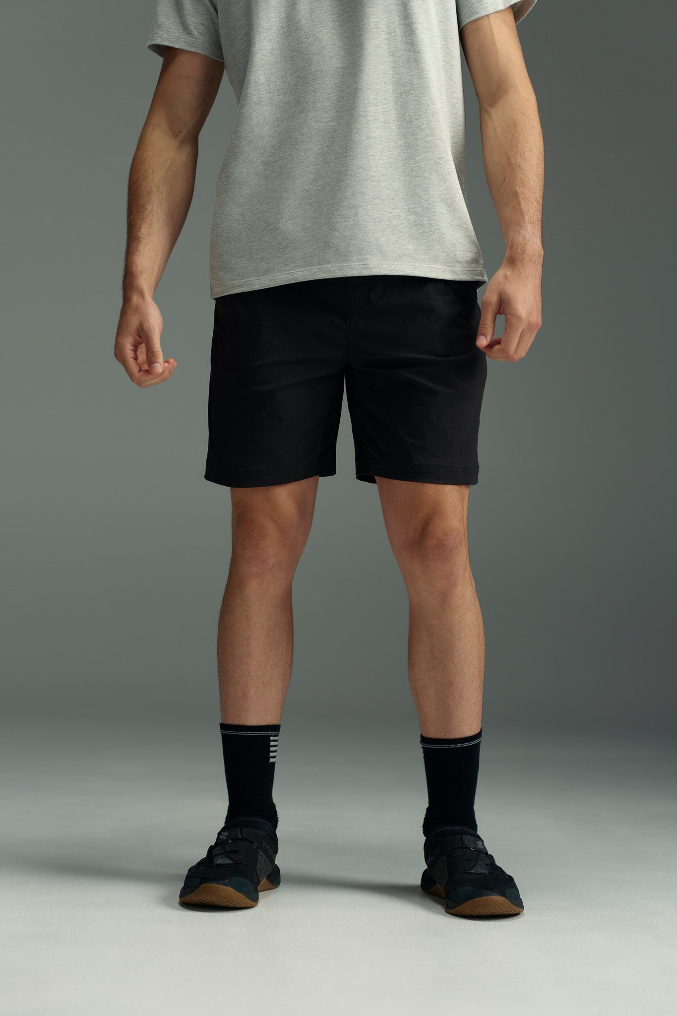 A person stands against a plain background, wearing a grey T-shirt and black WearEver Agile Shorts with an elastic waistband, paired with black socks with white stripes and black sneakers. Their face is not visible in the image.