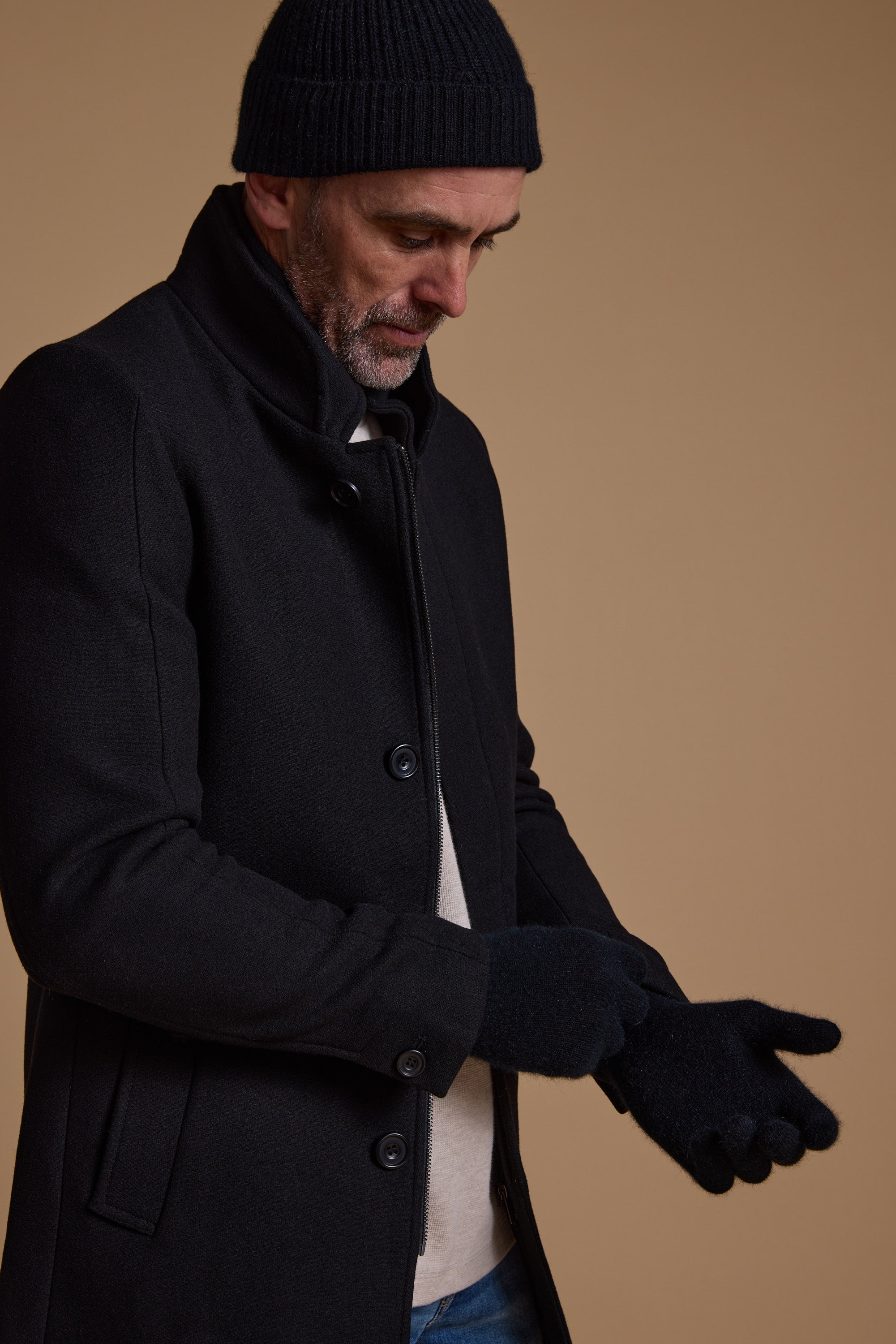 A man in a black coat and knit hat adjusts his NZ Made Perino Gloves by Woolyarns Perino, standing against a plain brown background.