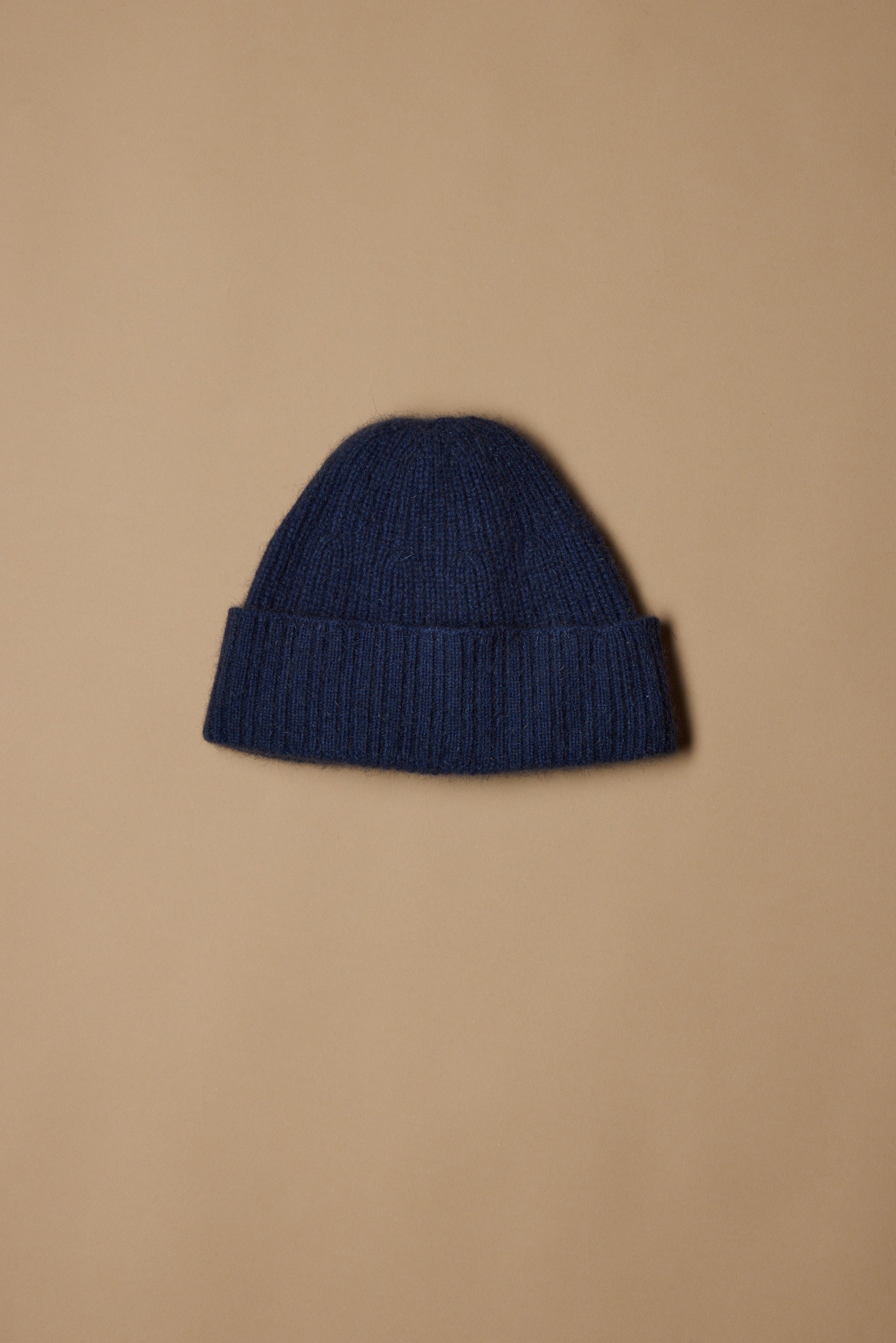 The Woolyarns Perino NZ Made Perino Beanie, crafted from a merino possum silk blend in dark blue with a folded brim, is shown against a light brown background.
