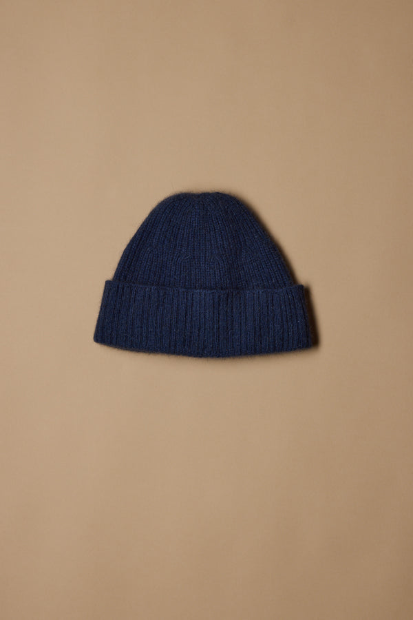 The Woolyarns Perino NZ Made Perino Beanie, crafted from a merino possum silk blend in dark blue with a folded brim, is shown against a light brown background.