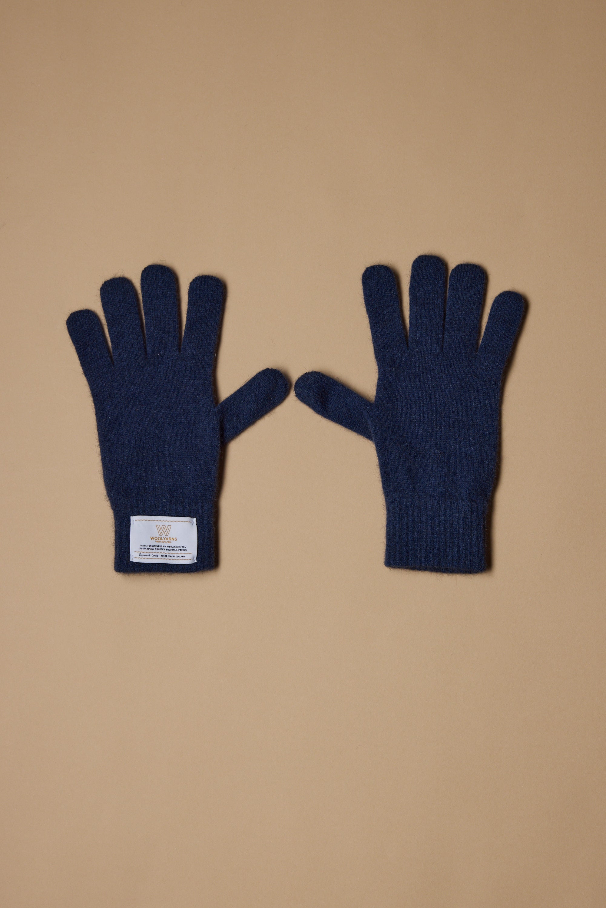 A pair of Woolyarns Perino NZ Made Perino Gloves in dark blue, crafted from a merino possum silk blend, is shown on a tan background. The left glove has a white rectangular wrist label; both gloves lie flat with fingers spread.