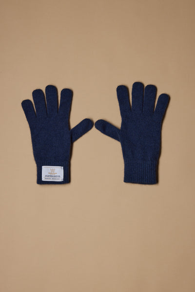 A pair of Woolyarns Perino NZ Made Perino Gloves in dark blue, crafted from a merino possum silk blend, is shown on a tan background. The left glove has a white rectangular wrist label; both gloves lie flat with fingers spread.