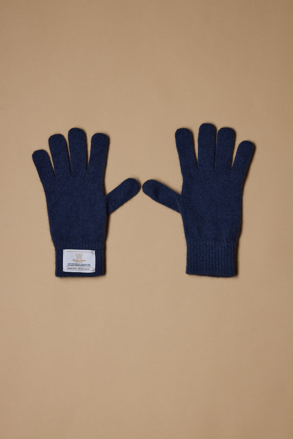 A pair of Woolyarns Perino NZ Made Perino Gloves in dark blue, crafted from a merino possum silk blend, is shown on a tan background. The left glove has a white rectangular wrist label; both gloves lie flat with fingers spread.