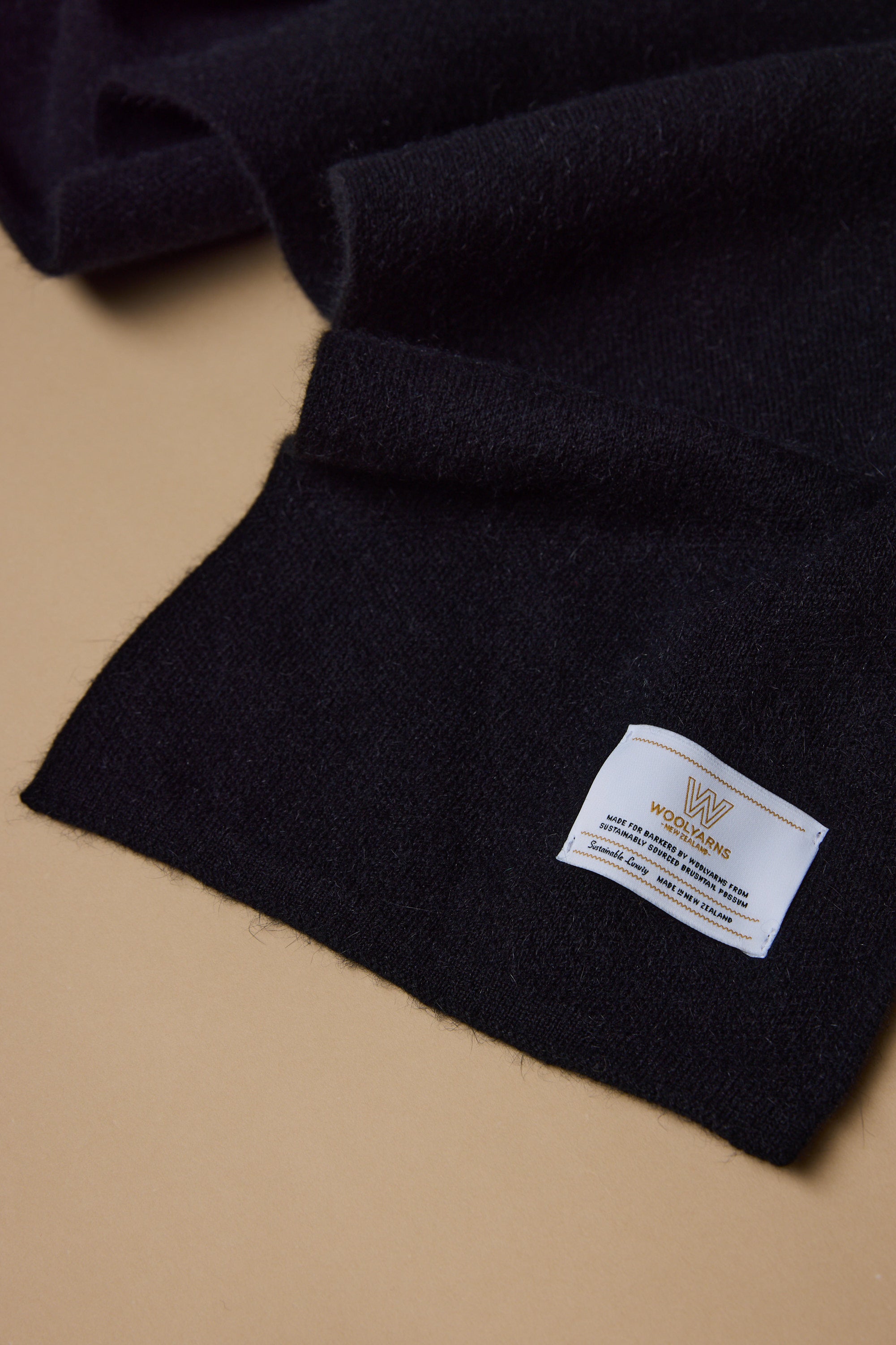 A close-up of the NZ Made Perino Scarf by Woolyarns Perino, in black wool and a luxurious merino possum silk blend, on a beige surface with a white label and gold WOOLMARK logo—crafted in New Zealand.