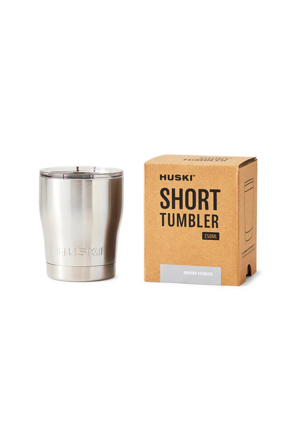 A Huski Short Tumbler 2.0 by Huski, made from stainless steel with a clear lid, is displayed next to packaging labeled “HUSKI SHORT TUMBLER 250ML—Brushed Stainless.” This vacuum-insulated tumbler keeps drinks at the perfect temperature.