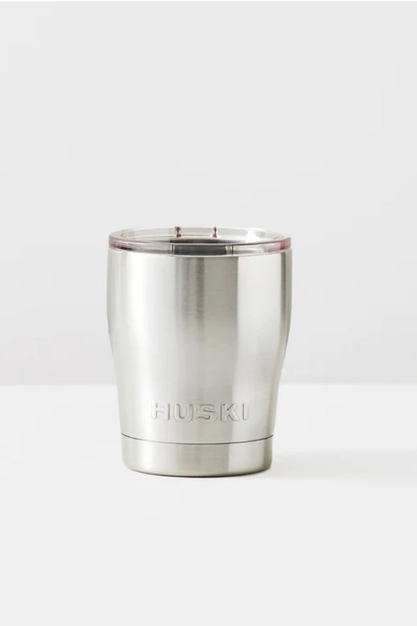 The Huski Short Tumbler 2.0 is a stainless steel, vacuum insulated tumbler with a clear plastic lid and the Huski logo engraved on the front. It sits on a white surface with a light grey background.