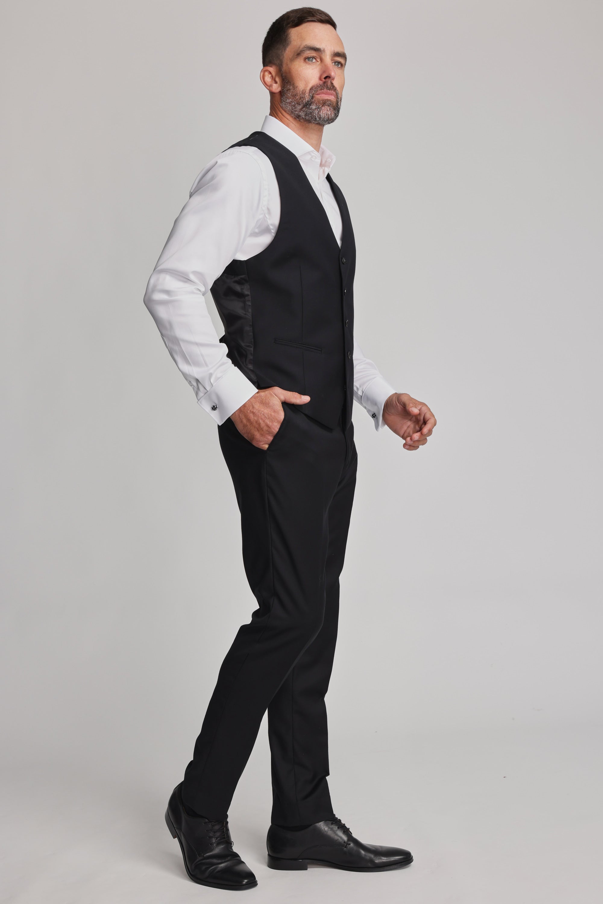 A man with short hair and a beard, dressed in a white shirt, Barkers Mayfair ZQrx Waistcoat, ZQrx Merino wool dress pants, and black shoes, stands in a side profile pose against a plain light background.