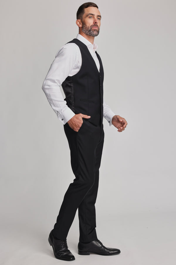 A man with short hair and a beard, dressed in a white shirt, Barkers Mayfair ZQrx Waistcoat, ZQrx Merino wool dress pants, and black shoes, stands in a side profile pose against a plain light background.