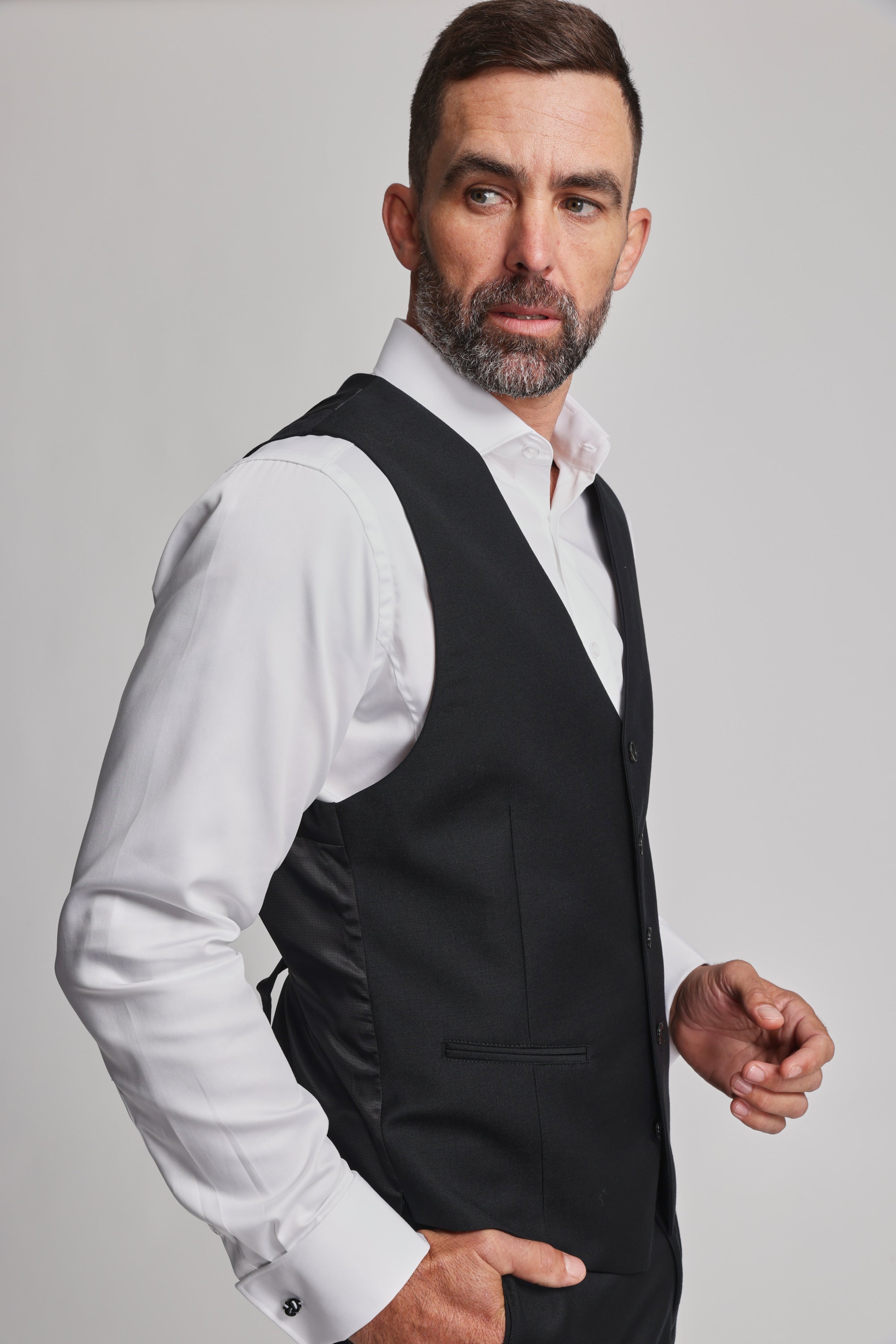 A man with short dark hair and a beard, wearing a white dress shirt and the Barkers Mayfair ZQrx Waistcoat, stands against a plain light background, looking to his left.