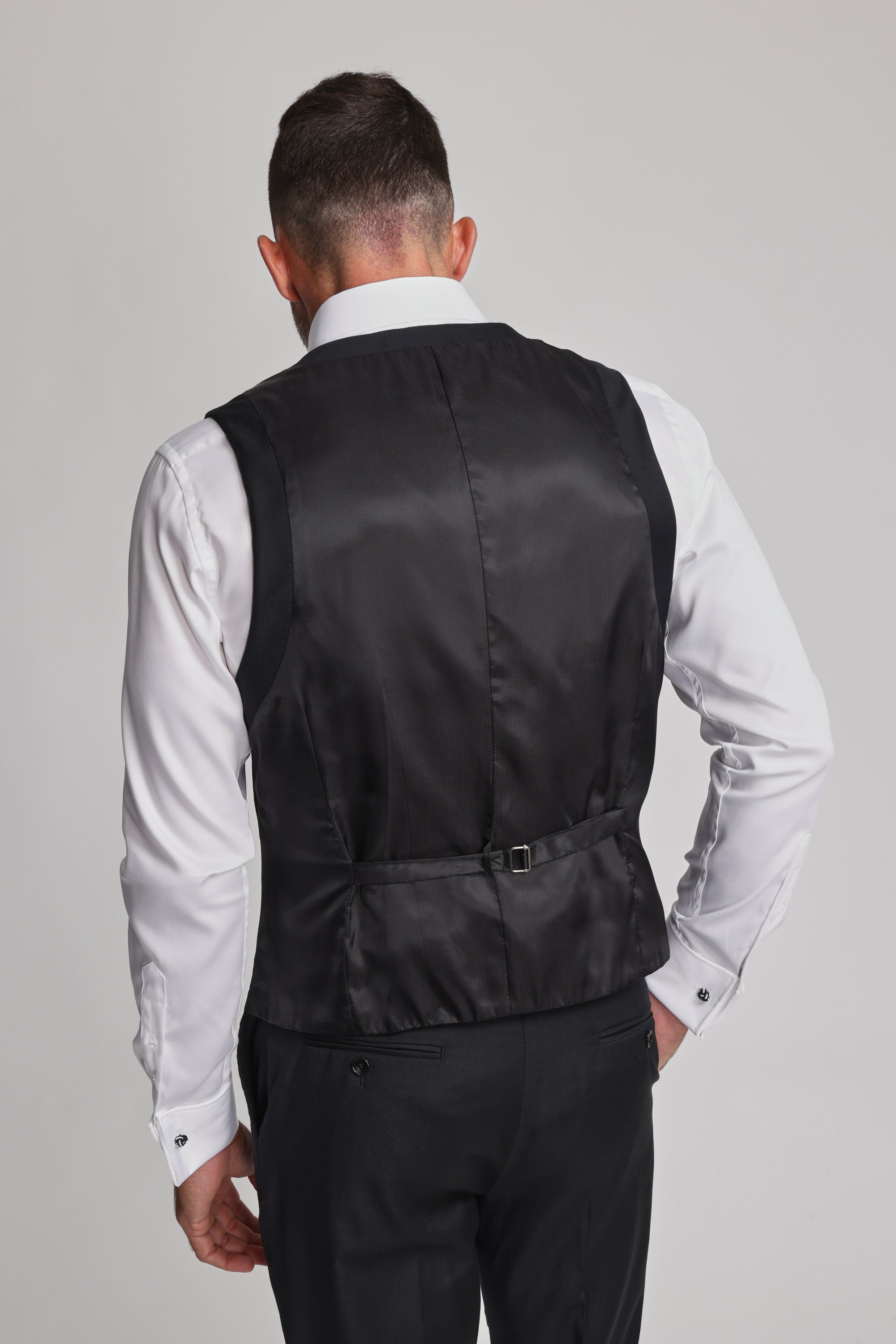 A man is seen from behind wearing the Barkers Mayfair ZQrx Waistcoat in black over a white shirt and black trousers. His right hand is on his hip, showing the vest’s adjustable strap. The background is plain and light-colored.