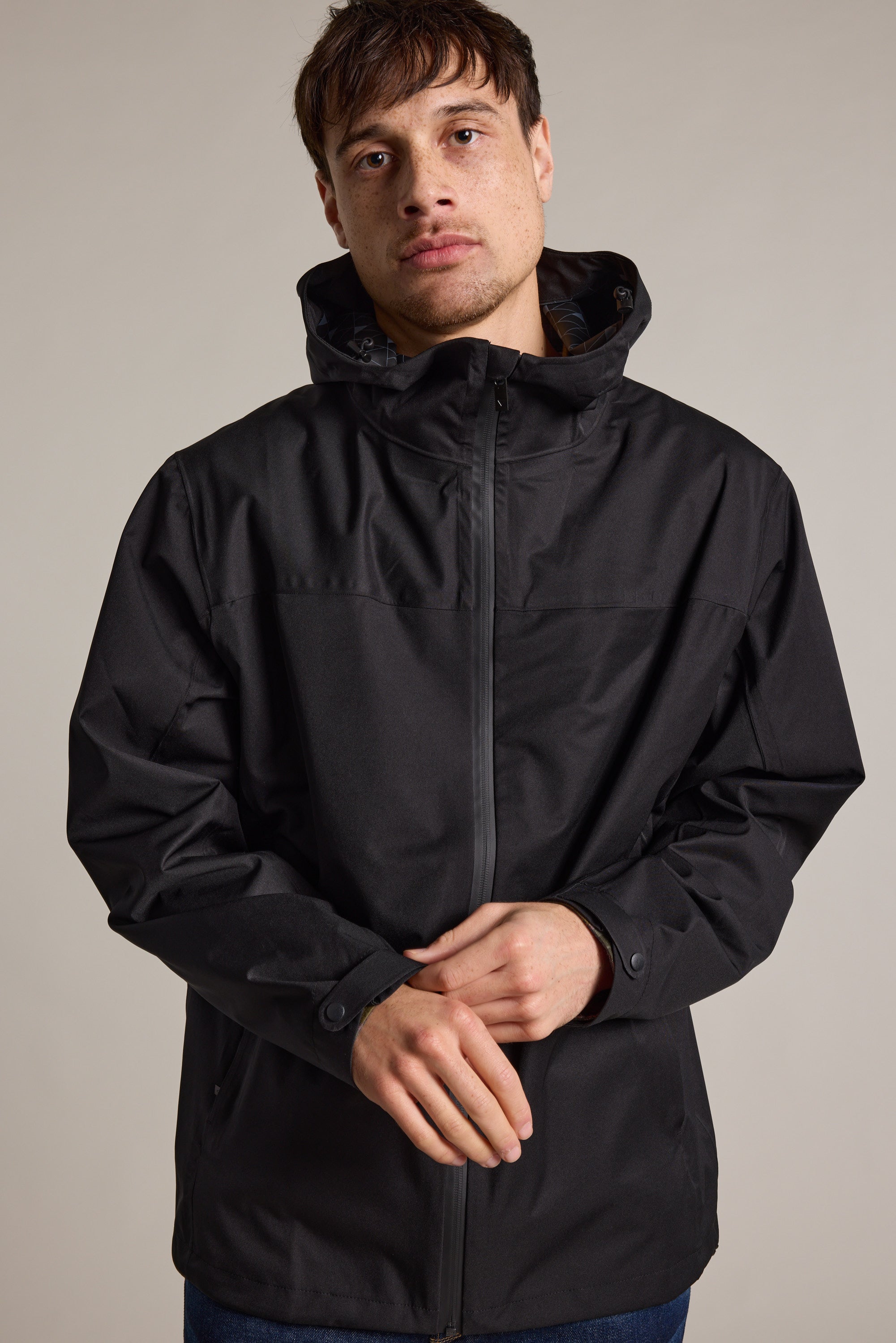 A person stands against a plain background, wearing the WearEver Warbler Outdoor Jacket in black, zipped to the neck, with hands loosely clasped in front and a neutral expression.