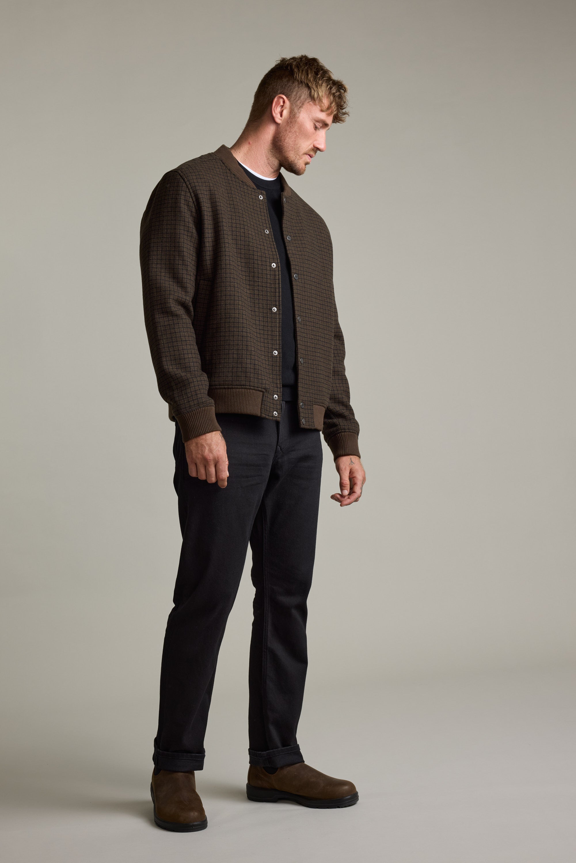 Man in Barkers Leland Wool Bomber Jacket with black shirt, pants, and brown boots looking down.
