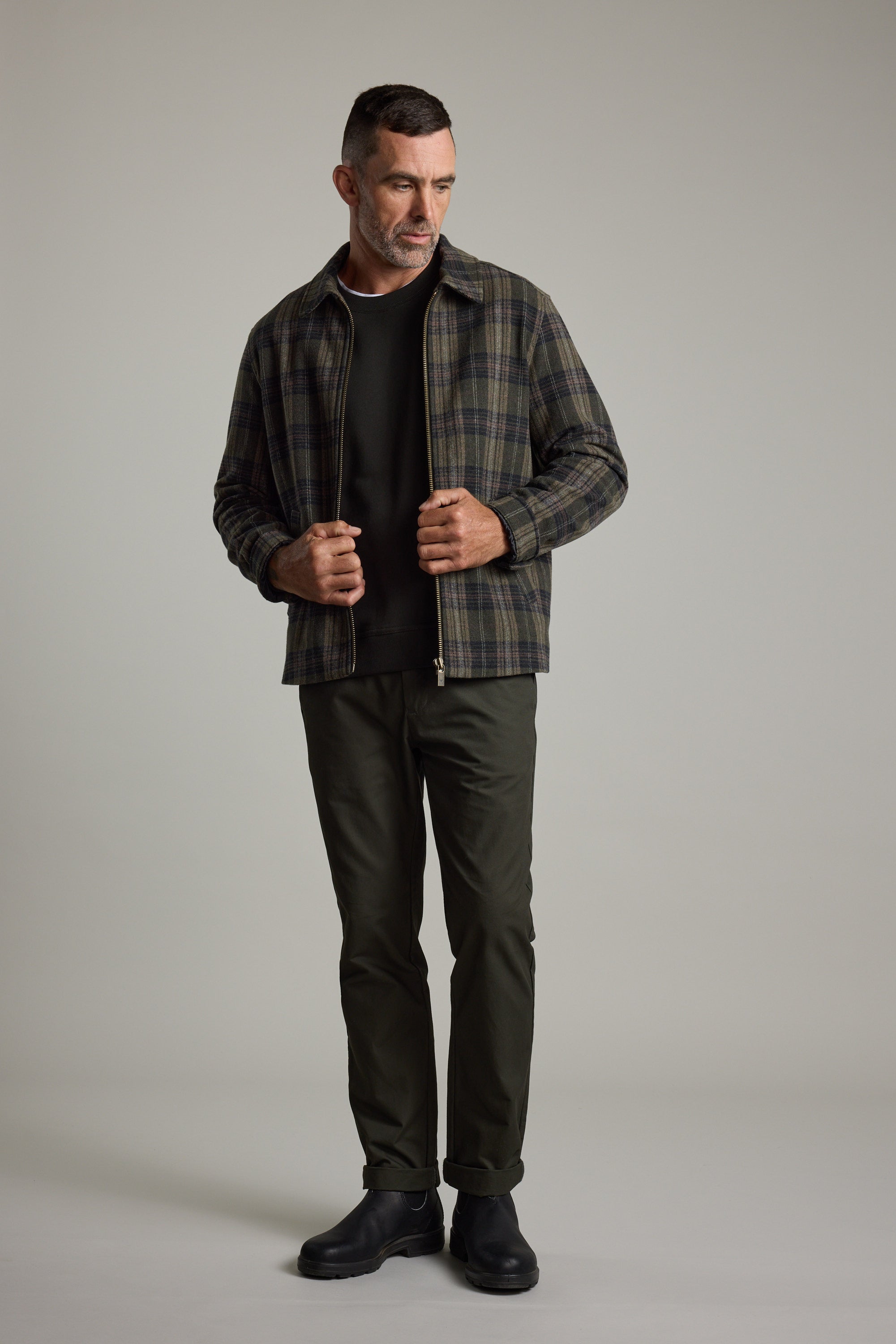 Man in Barkers Zermatt Wool Jacket, dark pants, and boots stands before a light gray background.