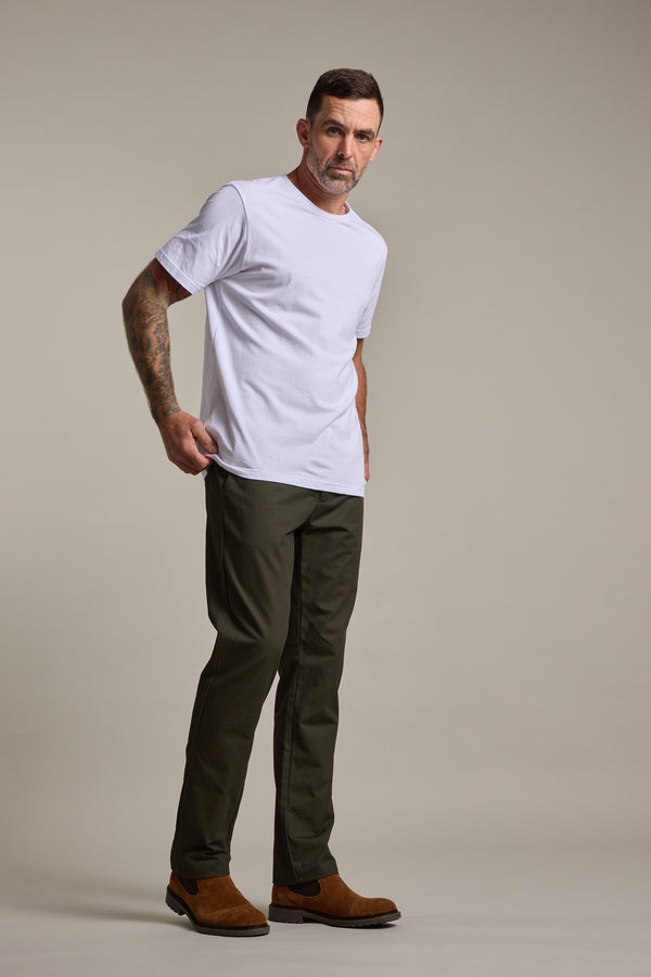 A man with short dark hair and a mustache, in a white t-shirt, Barkers Dundas Chino trousers in olive green, and brown shoes stands against a plain light backdrop, looking confidently at the camera.