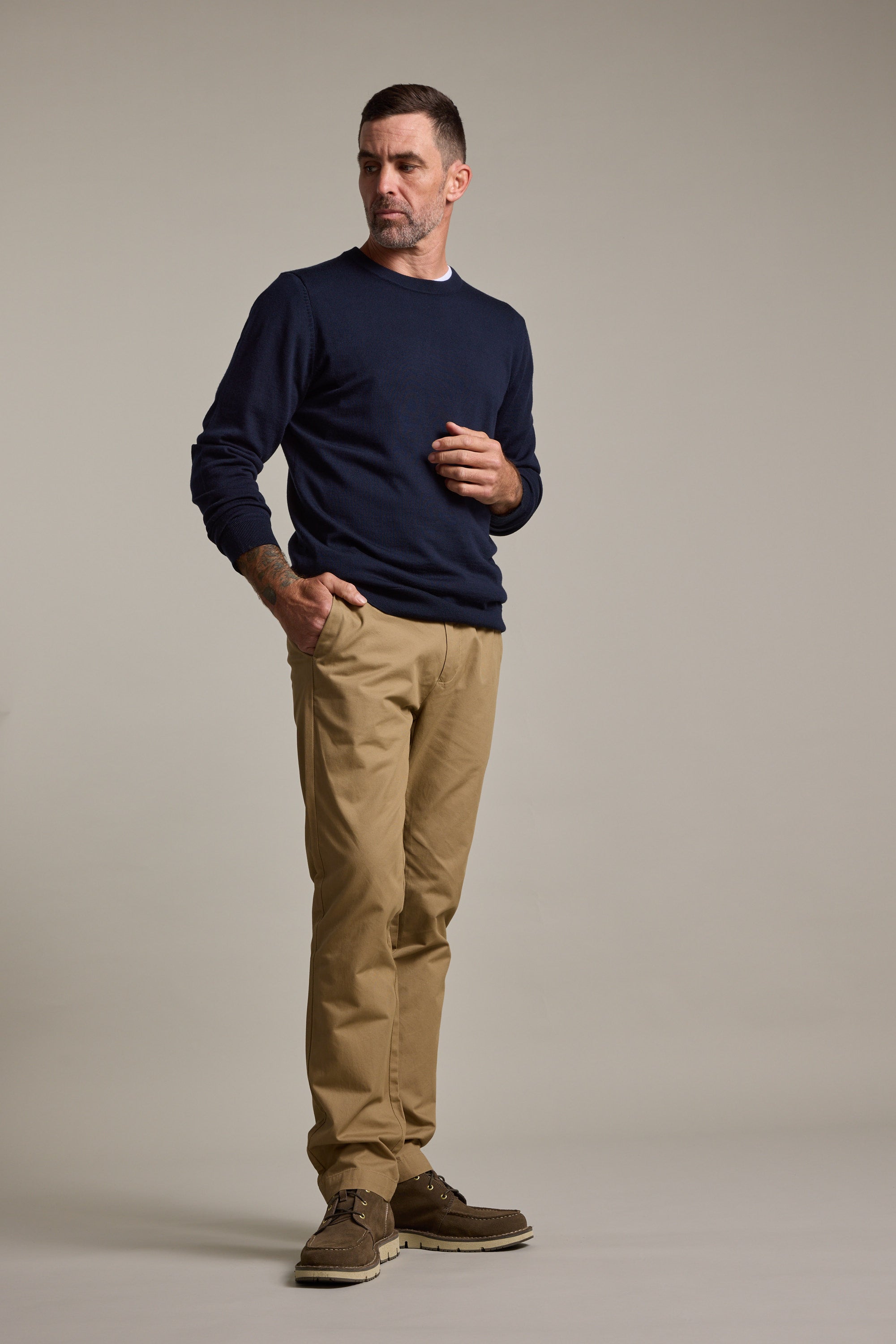 A man stands against a plain background, wearing Barkers Dundas Chino trousers, a navy sweater, and brown shoes, looking slightly to the side with one hand raised near his chest.