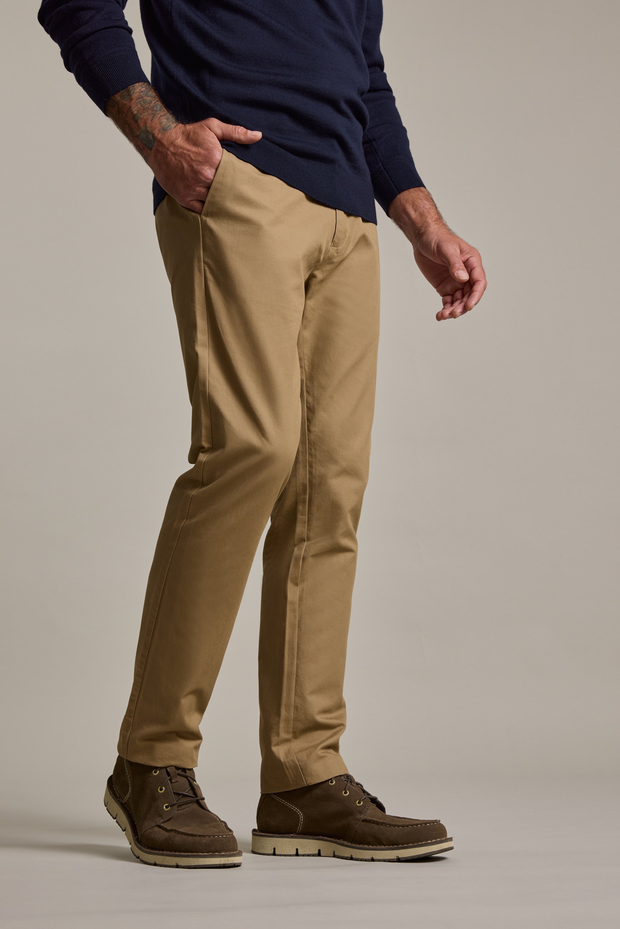 A person stands with one hand in their pocket, wearing Barkers Dundas Chino trousers, a navy sweater, and dark brown lace-up boots against a plain background.