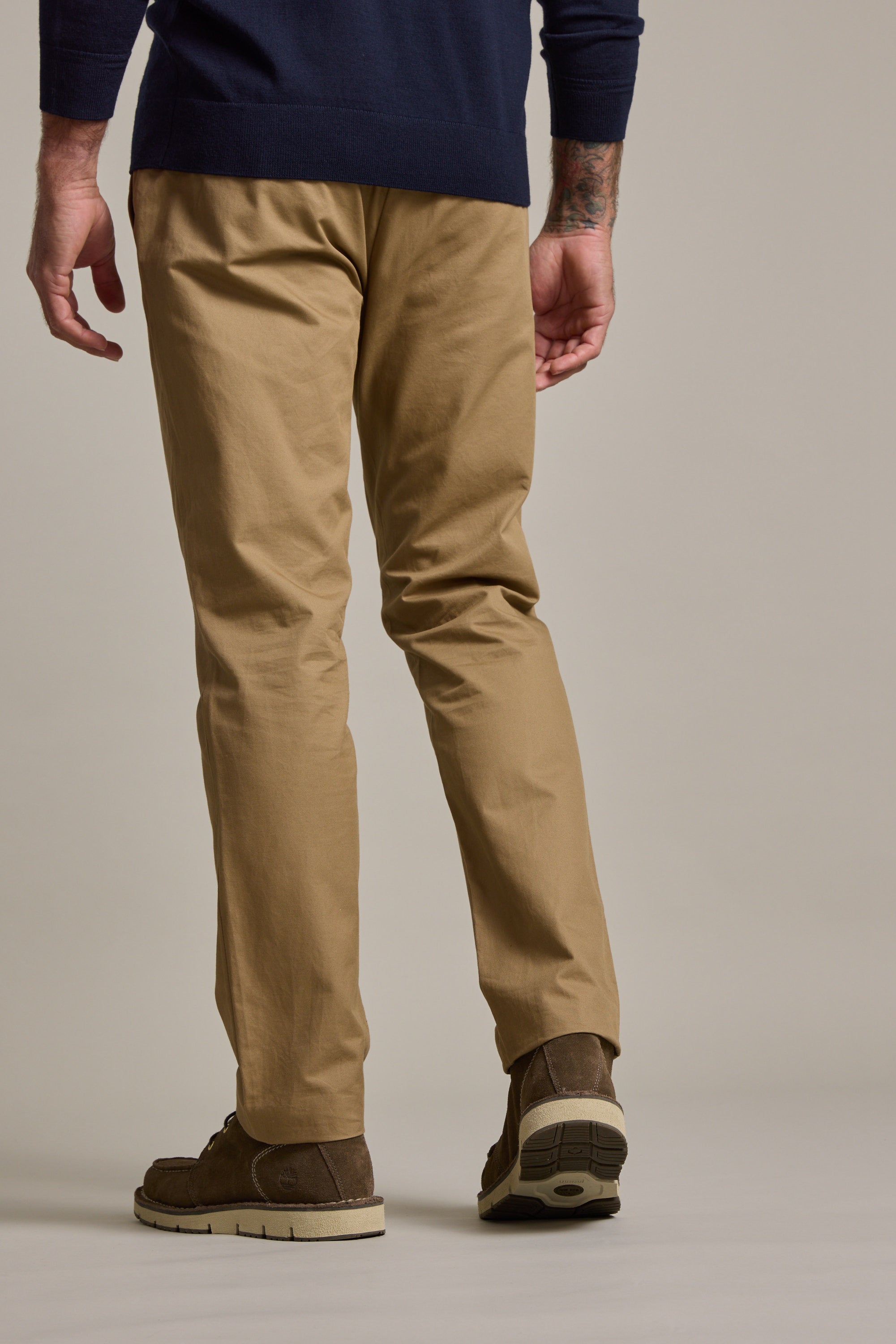 A person stands with their back to the camera, wearing Barkers Dundas Chino relaxed fit cotton twill pants, a dark blue long-sleeve shirt, and brown shoes against a plain neutral background.