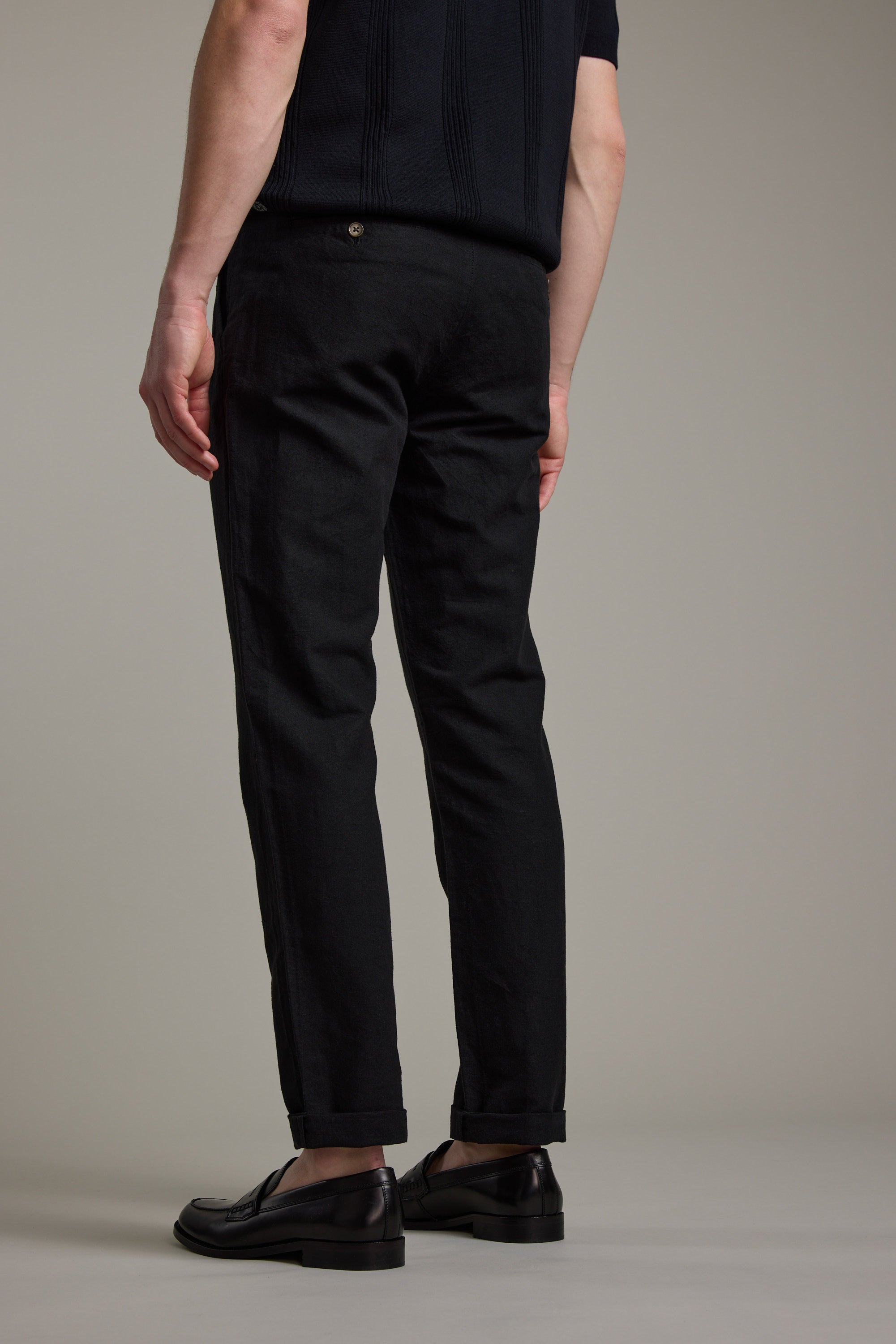 A person wearing Barkers’ Newman Linen Pant in black, paired with a short-sleeved black top and black dress shoes, stands facing away from the camera against a plain background.
