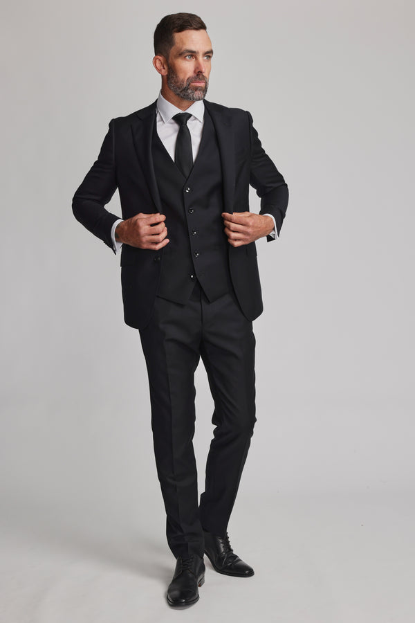 A man with short hair and a beard wears a Barkers Mayfair ZQrx Waistcoat as part of a black three-piece suit, paired with a white dress shirt and black tie. He stands confidently against a plain light background, adjusting his jacket.