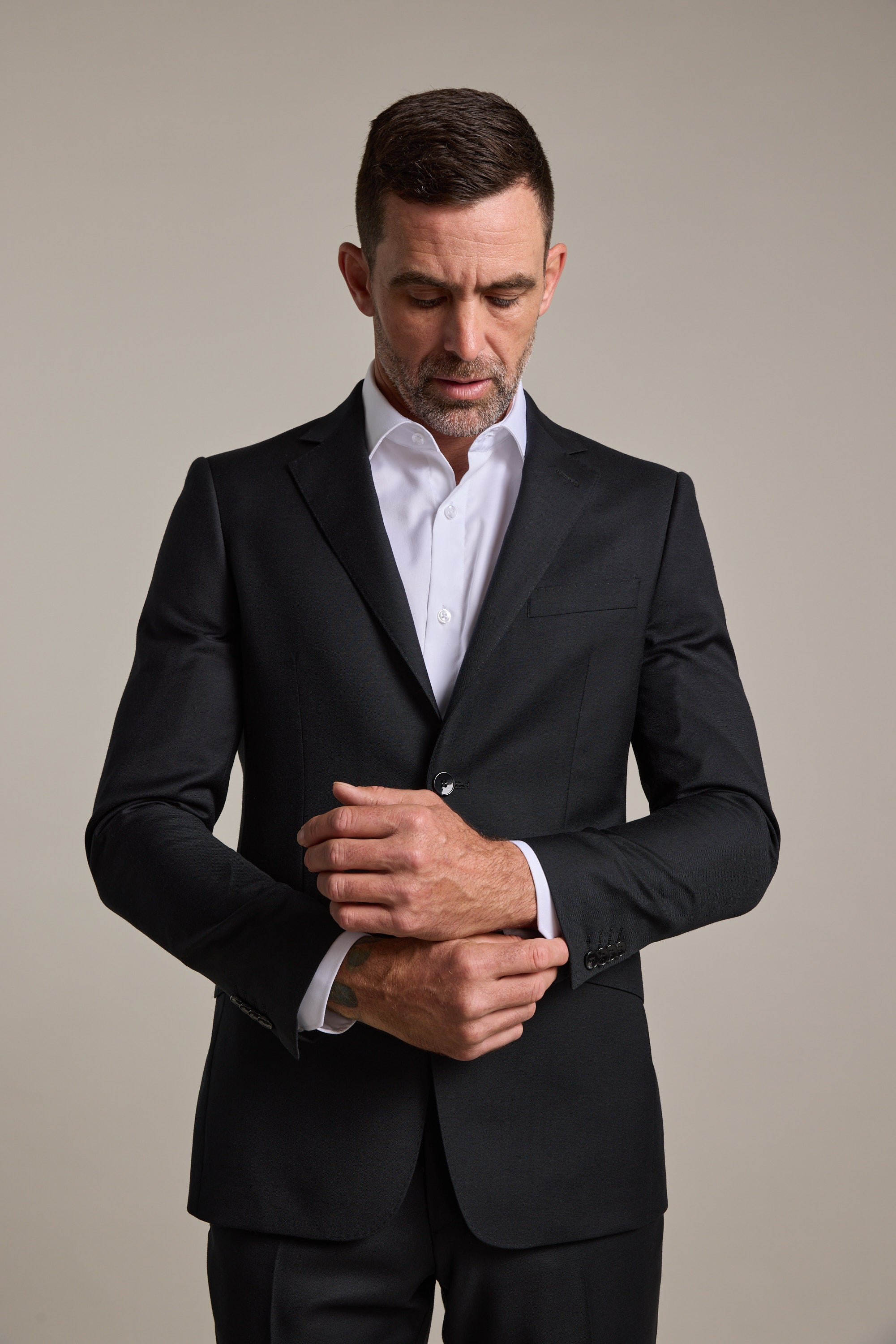 A man wearing the Barkers Mayfair ZQrx Suit Jacket in black merino wool and a white dress shirt stands against a plain background, looking down as he adjusts his cuff.