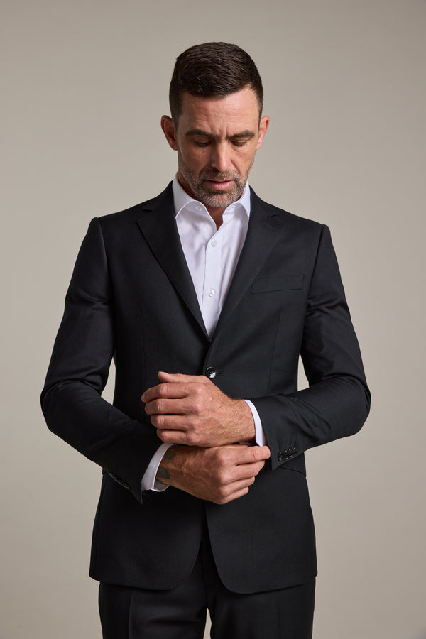 A man wearing the Barkers Mayfair ZQrx Suit Jacket in black merino wool and a white dress shirt stands against a plain background, looking down as he adjusts his cuff.