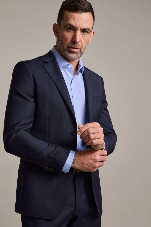 A man with short brown hair and a trimmed beard wears the Barkers Mayfair ZQ Suit Jacket in navy over a light blue dress shirt. He buttons his jacket and looks slightly to the side against a plain, light background.