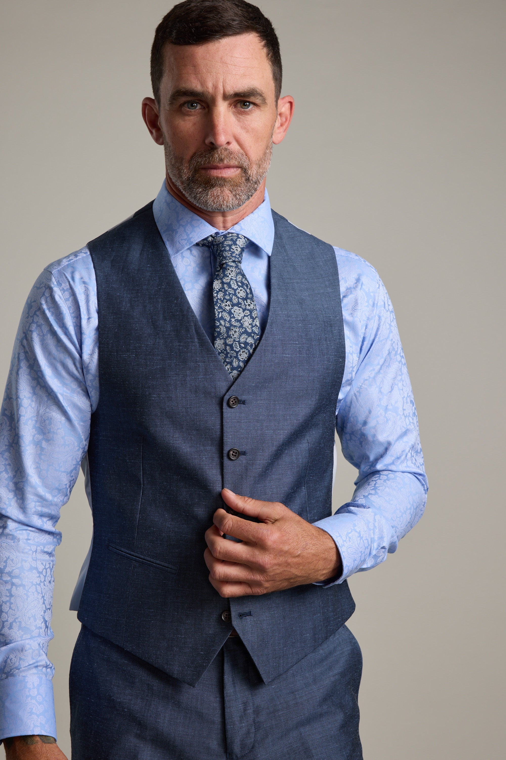 A man with short dark hair and a trimmed beard wears a blue three-piece suit with the Barkers Charles Texture Waistcoat, an adjustable linen wool piece. He pairs it with a light blue patterned shirt, floral tie, and adjusts his vest while looking serious.