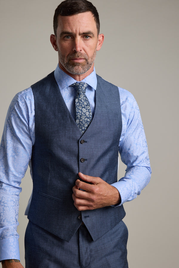 A man with short dark hair and a trimmed beard wears a blue three-piece suit with the Barkers Charles Texture Waistcoat, an adjustable linen wool piece. He pairs it with a light blue patterned shirt, floral tie, and adjusts his vest while looking serious.